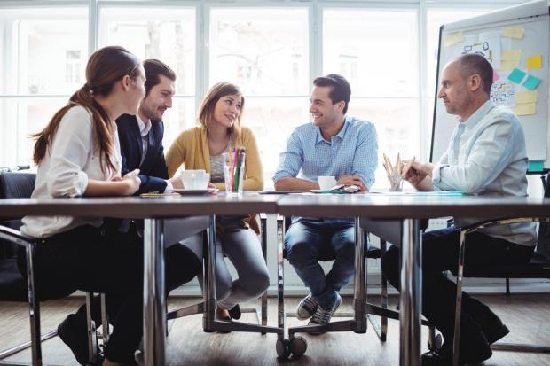Image of colleagues chatting at a table in front of a whiteboard